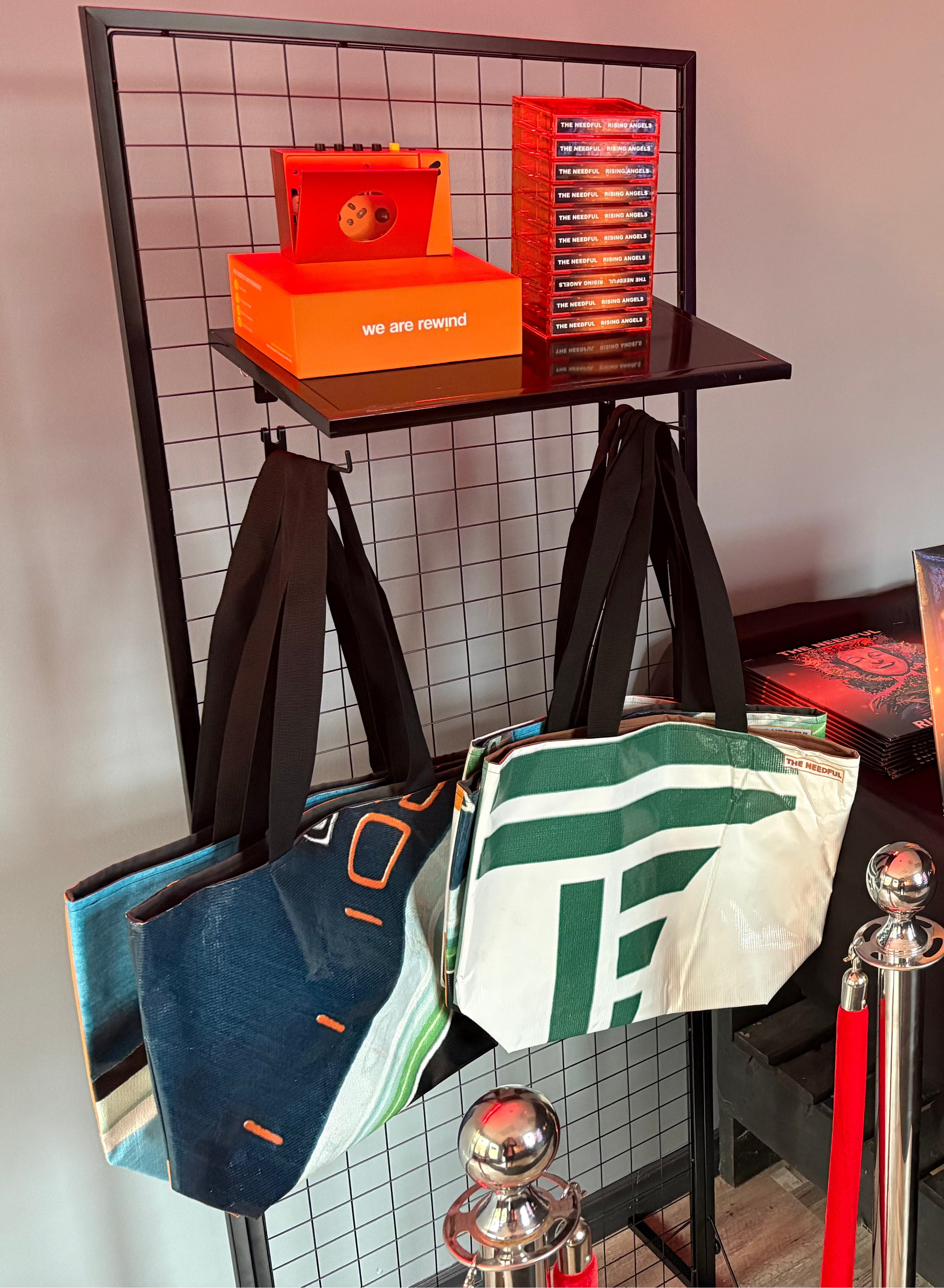 A product display booth for 'The Needful' featuring merchandise including tote bags, t-shirts, casette, album, and orange branded boxes on a black wire rack and a black-clothed table.