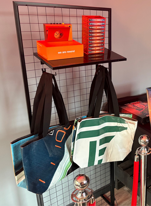A product display booth for 'The Needful' featuring merchandise including tote bags, t-shirts, casette, album, and orange branded boxes on a black wire rack and a black-clothed table.