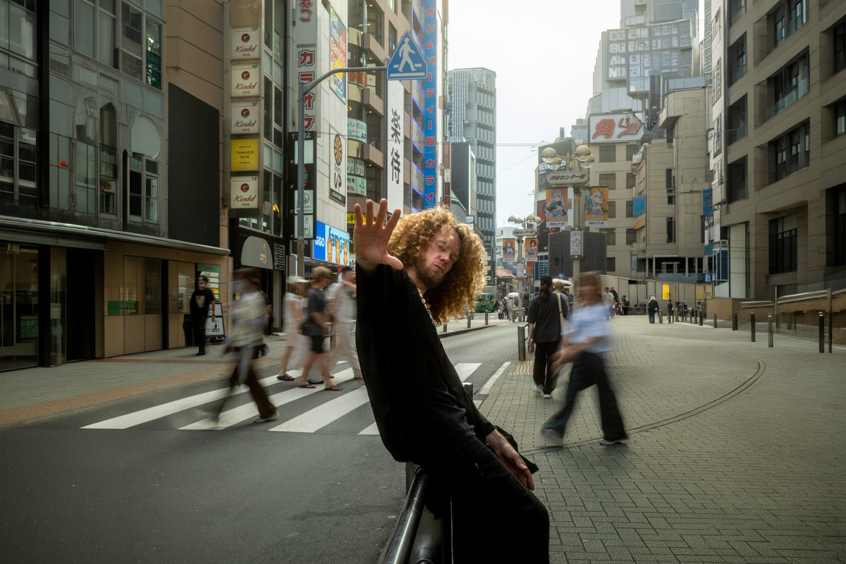 A person with curly hair wearing a black outfit leans against a metal railing on a city street, waving at the camera. Pedestrians walk in the background of an urban street scene with tall buildings and signs.
