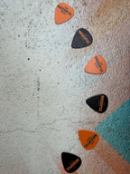 Several guitar picks arranged diagonally on a textured light gray surface. The picks are orange and black with the text 'NEEDFUL MUSIC' printed on them.