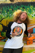 A man with curly blonde hair stands in front of a colorful mural, wearing a black-and-white raglan shirt with a circular graphic that reads 'The Needful Rising Angels.' Several branded orange items, a blue bag, and other items are arranged on a table beside him.