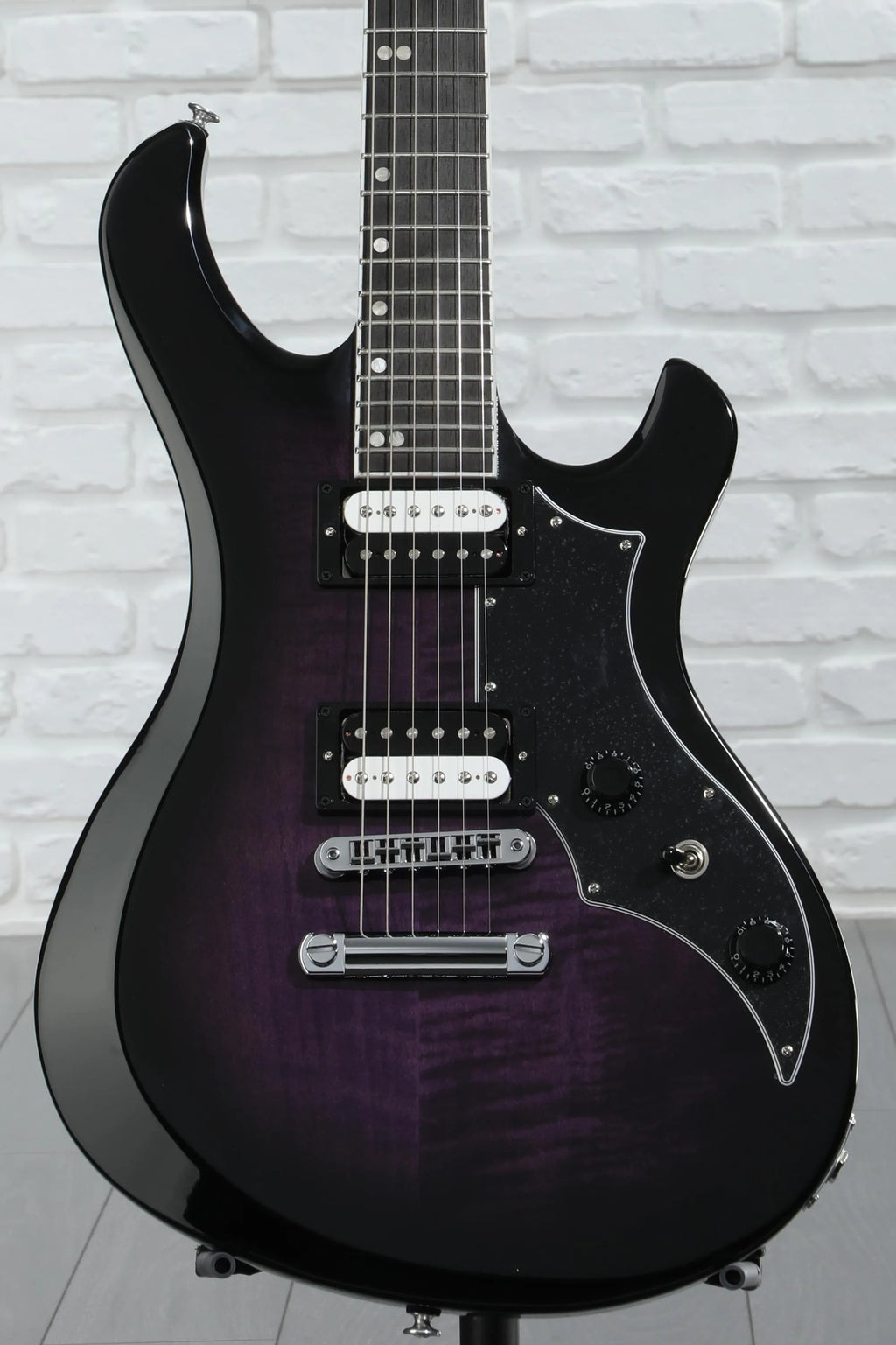 Black electric guitar with a purple tint against a light gray brick wall.