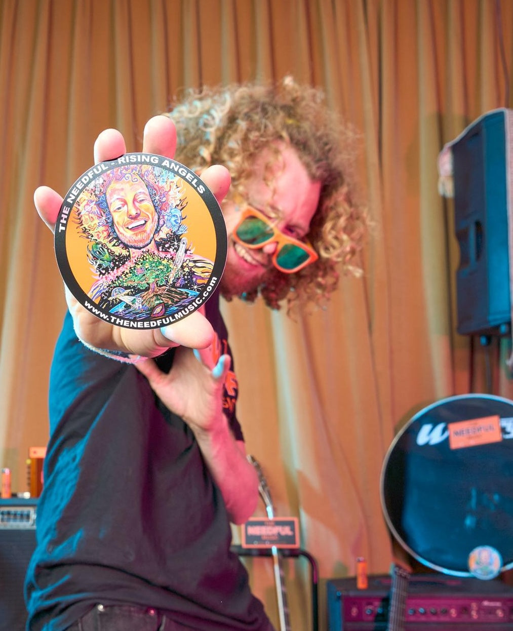 Glenn holding a colorful round sticker with 'The Needful Rising Angels' album branding in front of a stage setup.