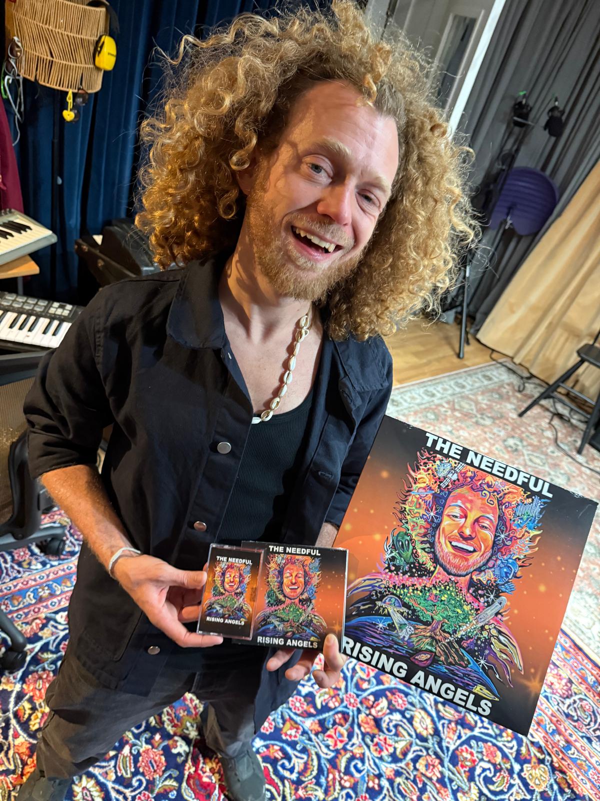 Glenn holding 'The Needful Rising Angels' album and merchandise in a studio setting