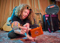 Glenn of The Needful holding a colorful cassette player and a round object with Rising Angels album design, surrounded by musical instruments.
