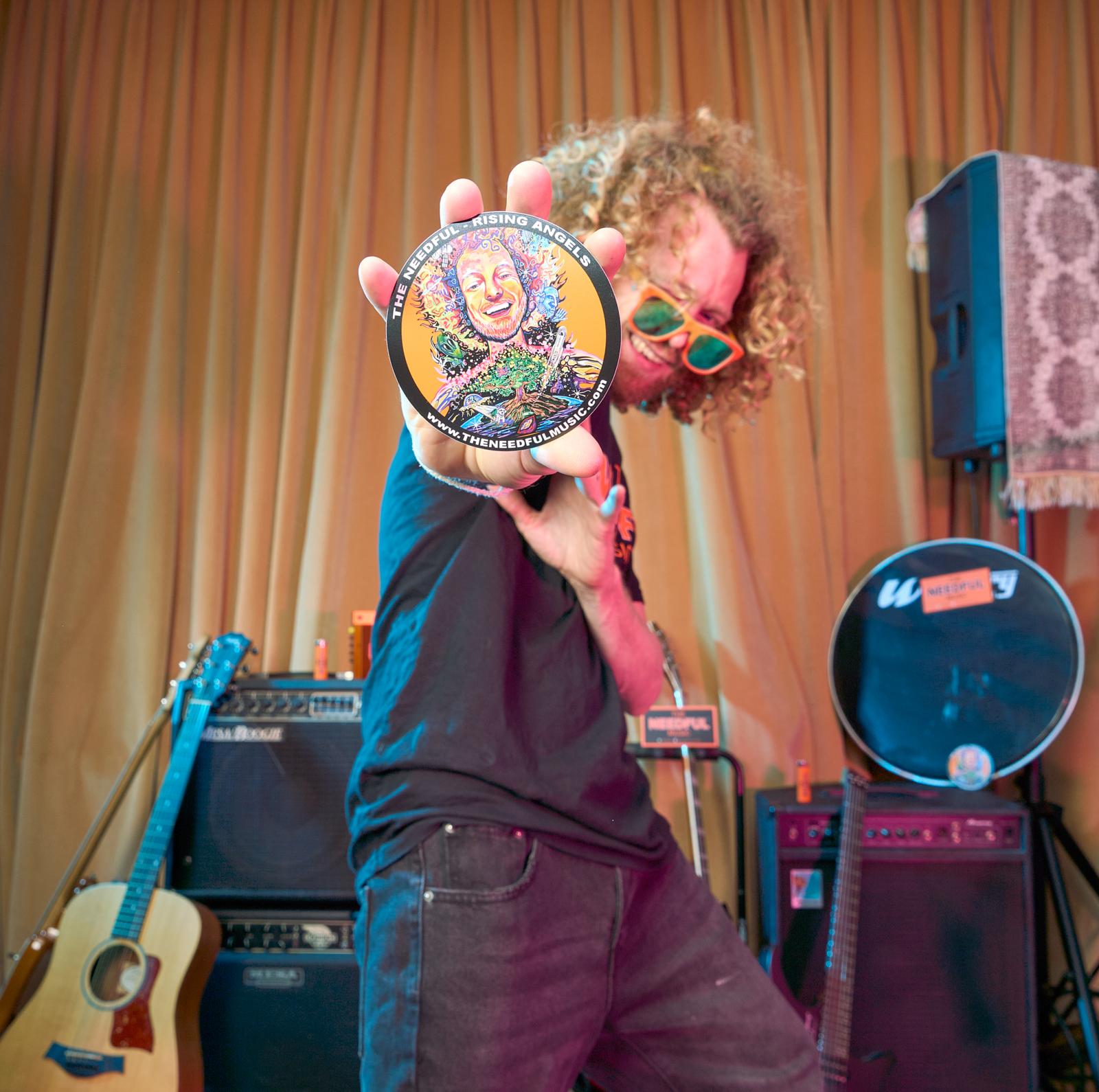 Person holding a colorful circular sticker in front of a musical setup with instruments and equipment.