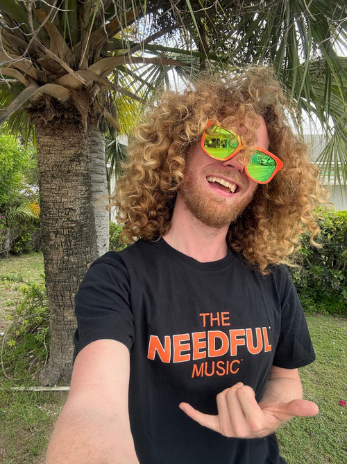 Glenn, singer of The Needful wearing orange wayfarer-style sunglasses in The Needful tshirt in a tropical location.