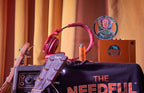 Music-related items including a guitar, headphones, and a 'The Needful' shirt on a table with a warm-toned curtain background.