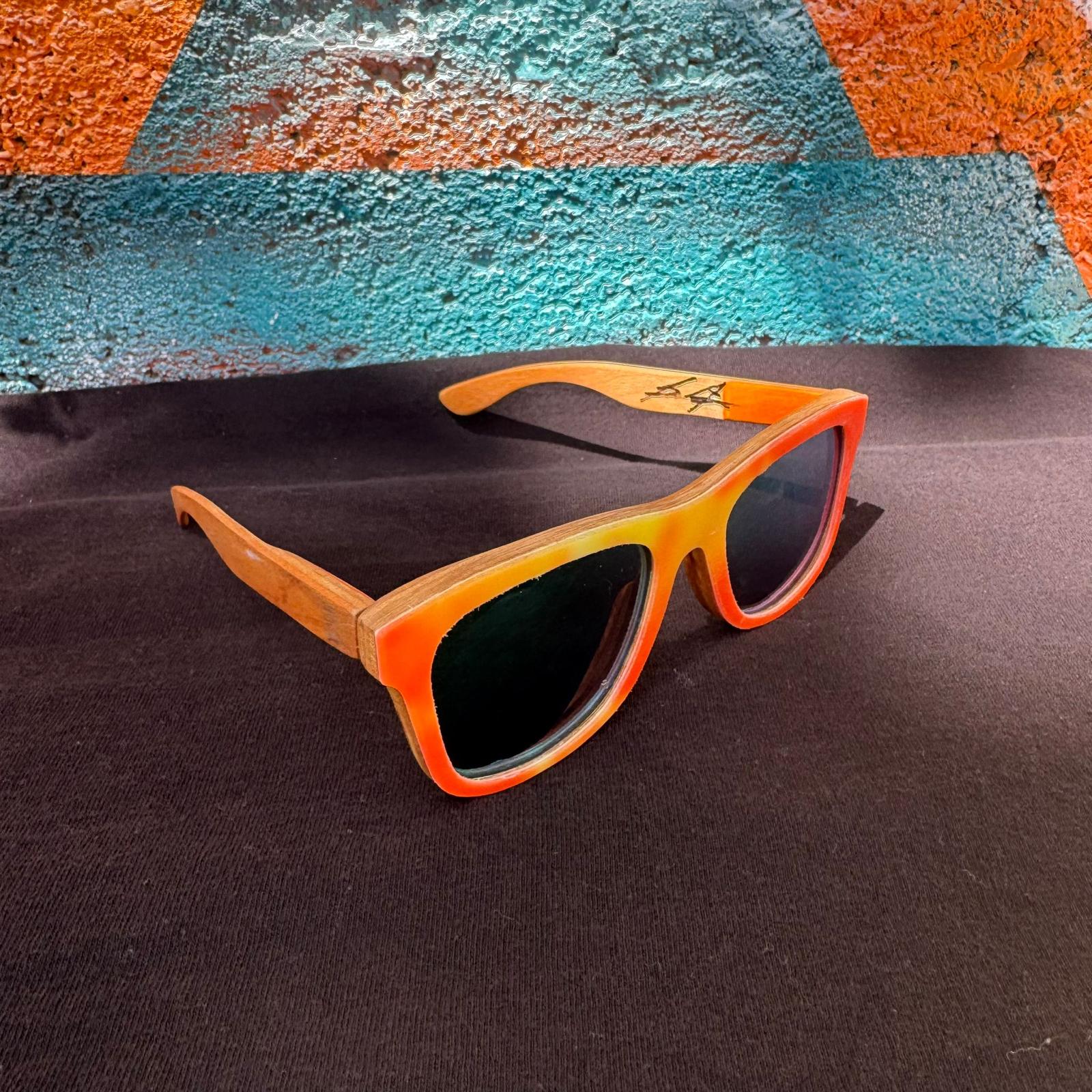Wooden-framed sunglasses with a vibrant orange-to-yellow gradient finish and dark lenses, resting on a dark textured surface with a colorful painted background.