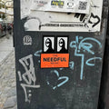Sticker-covered black surface with various stickers, including 'The Needful Music' from an indie band in Canada.