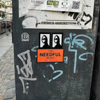 Sticker-covered black surface with various stickers, including 'The Needful Music' from an indie band in Canada.