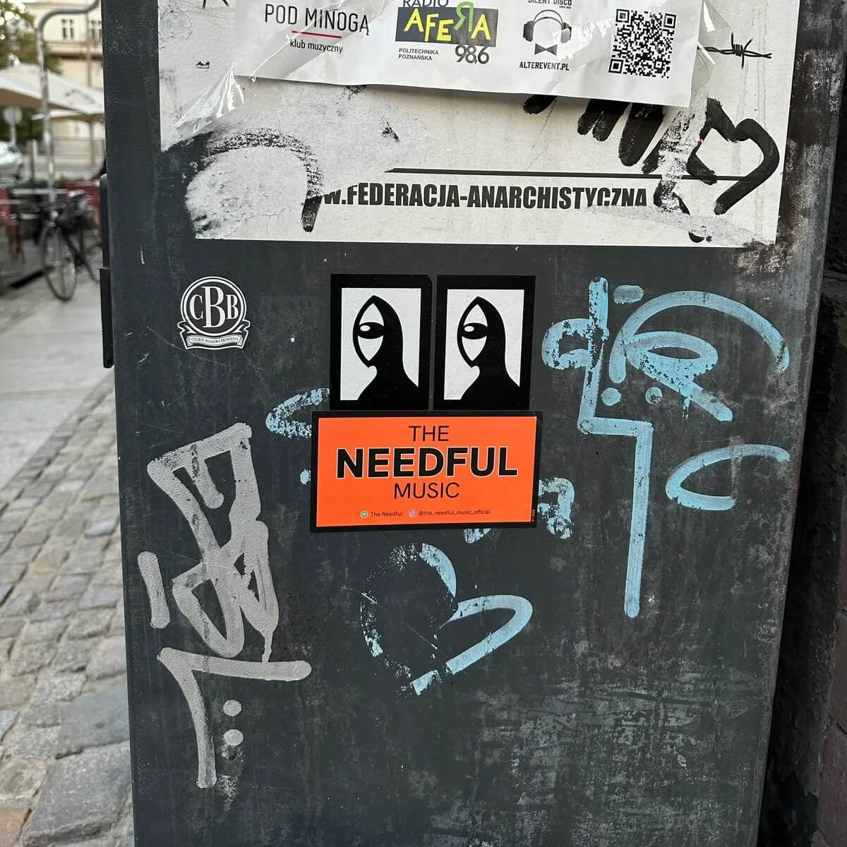 Sticker-covered black surface with various stickers, including 'The Needful Music' from an indie band in Canada.