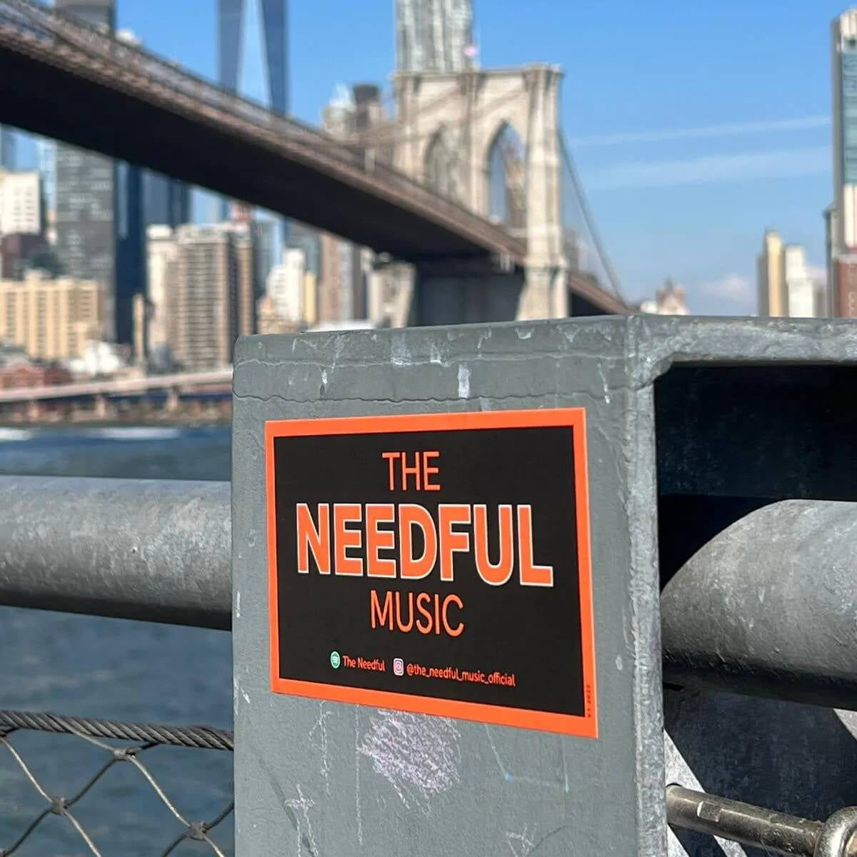 Sticker for 'The Needful Music' on a metal railing with the Brooklyn Bridge and city skyline in the background.