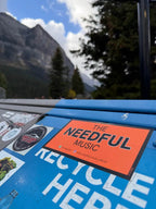 Sticker for 'The Needful Music' on a blue surface with mountains in the background at Lake Louise Canada
