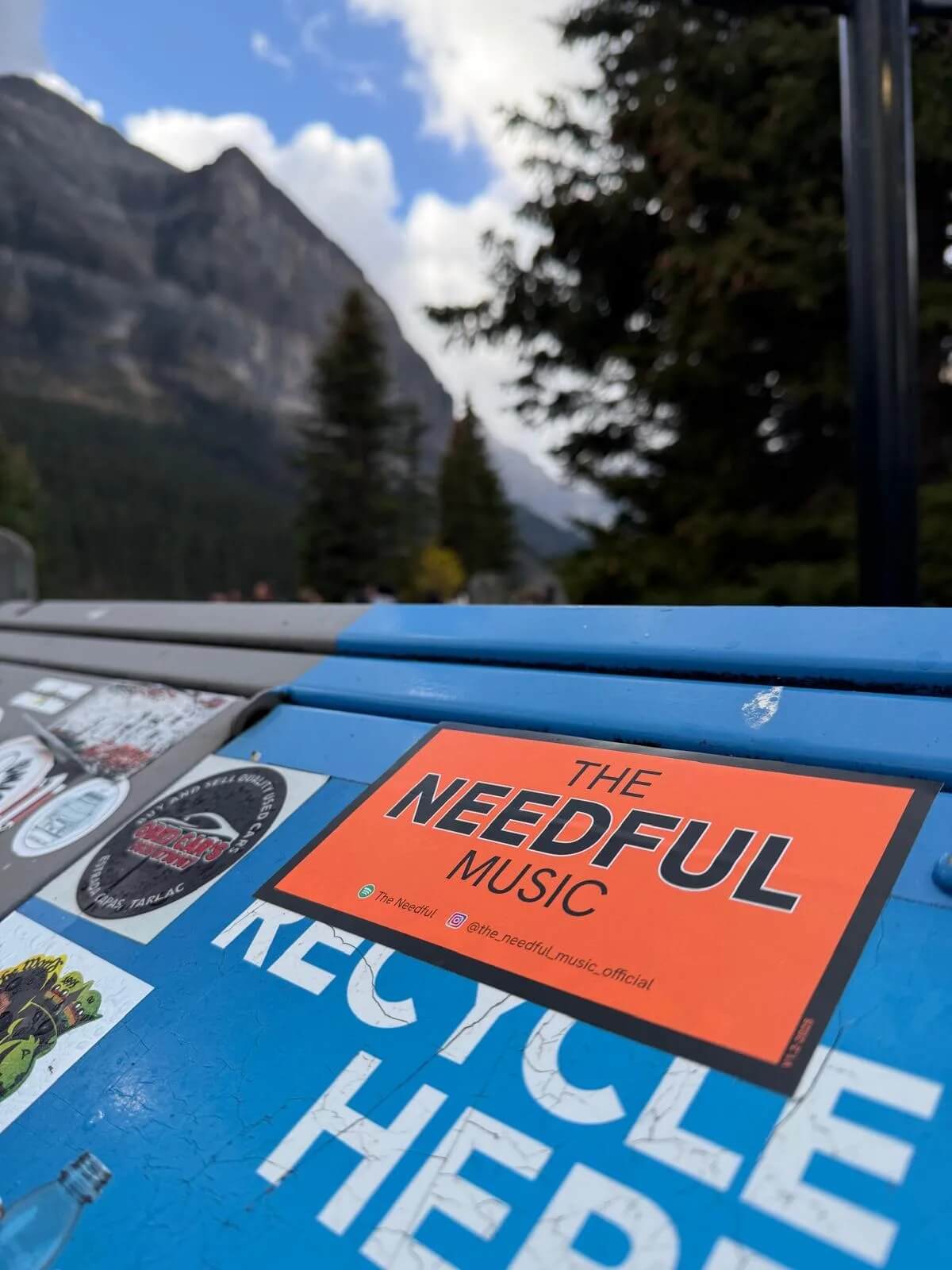 Sticker for 'The Needful Music' on a blue surface with mountains in the background at Lake Louise Canada