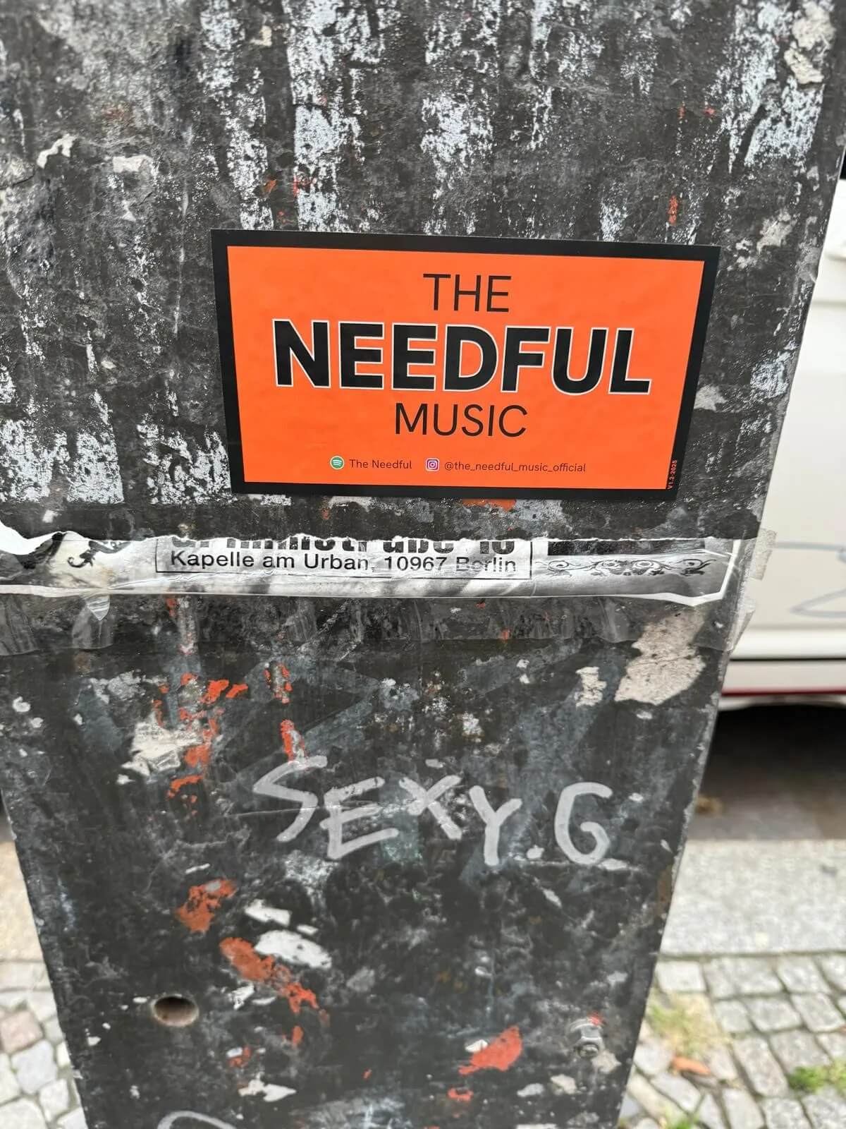 Orange sticker with 'The Needful Music' text on a textured surface with graffiti.
