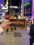 Hand holding a 'The Needful Music' sticker in a city street at night.