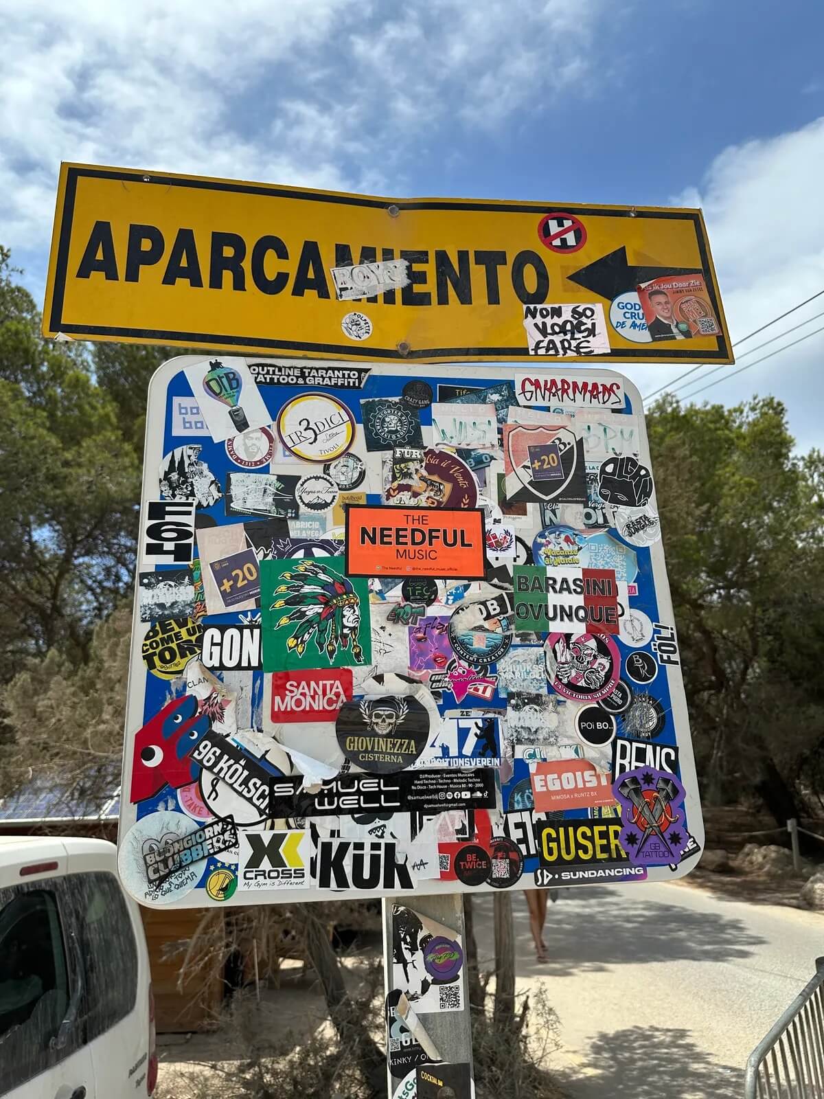 Signboard with various stickers, including the orange The Needful sticker in the middle and a yellow sign above it on a road in south america.
