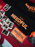 Black t-shirt with 'The Needful Music' branding on a red background with stickers and polaroid pictures of the band.