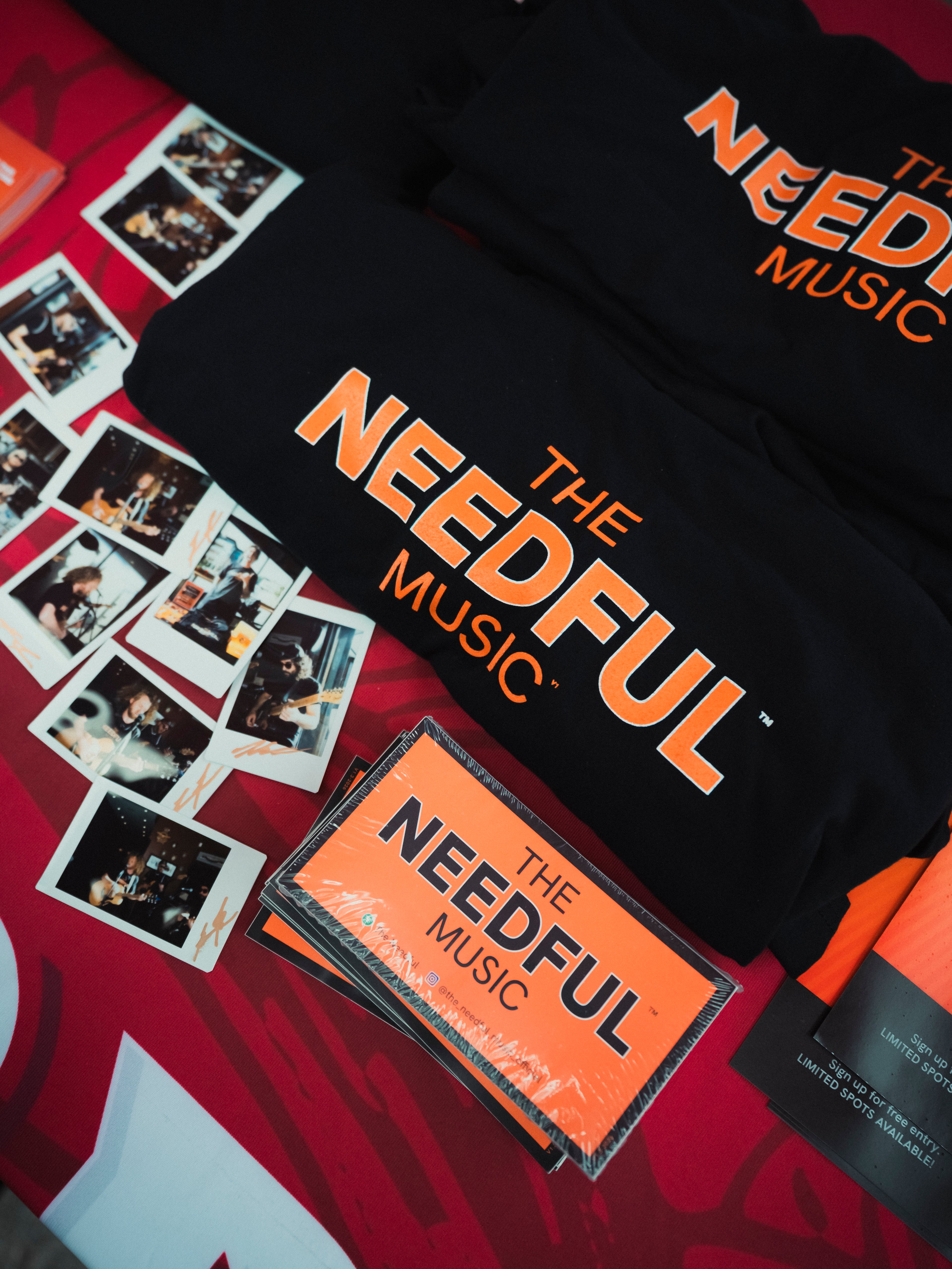 Black t-shirt with 'The Needful Music' branding on a red background with stickers and polaroid pictures of the band.