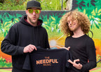 Two men, Travis and Glenn, holding a 'The Needful Music' t-shirt in front of a colorful mural and wearing the orange sunglasses.