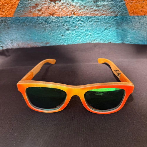 A pair of wayfair style sunglasses with a vibrant orange and yellow gradient frame and dark green lenses, resting on a dark textured surface with a colorful blue and orange background.